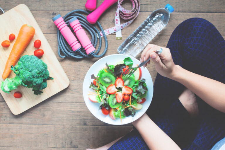 Mujer sentada en posición de loto con un bowl de comida saludable, rodeada de verduras, agua y cuerda de ejercicio, representando hábitos saludables diarios.